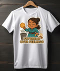 White t-shirt with a cartoon character holding frybread and text 'Frybread Over Feelings' on a wooden background.
